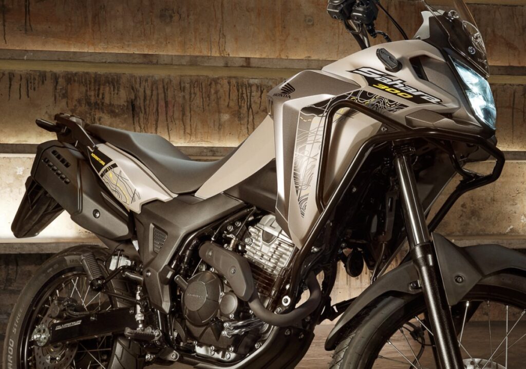 Imagem da nova Honda XRE 300 Sahara em destaque. Moto cinza com rodas grandes raiadas pintadas de preto, suspensão longa e perfil aventureiro. Lançamento para o mercado brasileiro.