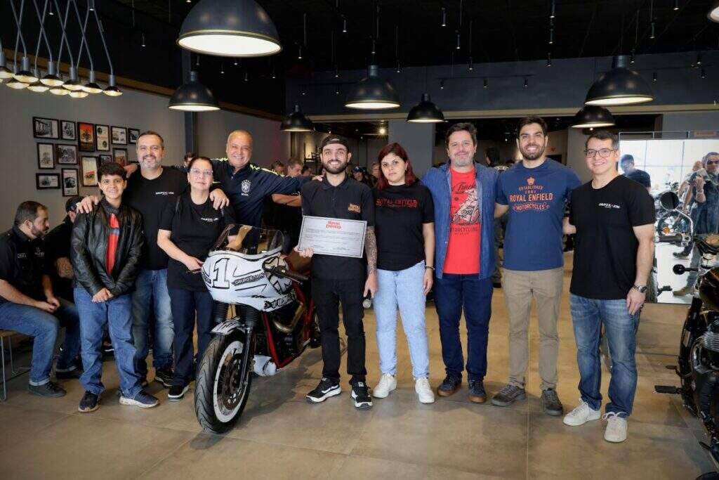 Um grupo de pessoas, incluindo funcionários da Royal Enfield e do Grupo Raion, posando para uma foto dentro da concessionária. No centro da imagem, há uma motocicleta customizada em destaque, e algumas pessoas seguram um certificado ou placa.